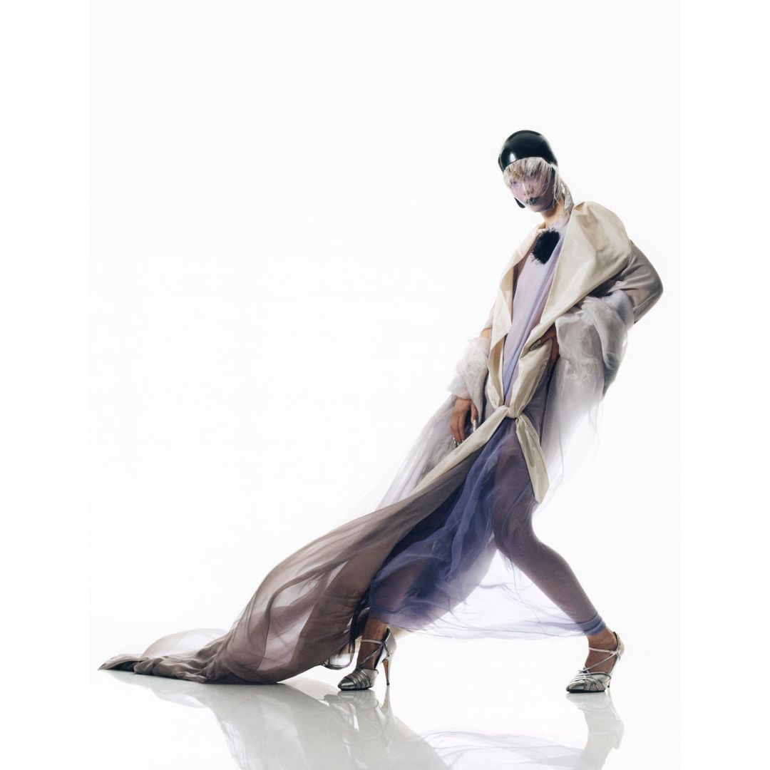 A person poses in a light coat and sheer flowing fabric against a white backdrop, showcasing a high-fashion editorial style.