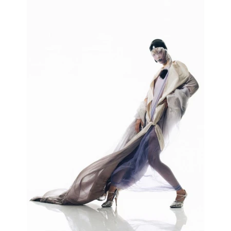 A person poses in a light coat and sheer flowing fabric against a white backdrop, showcasing a high-fashion editorial style.