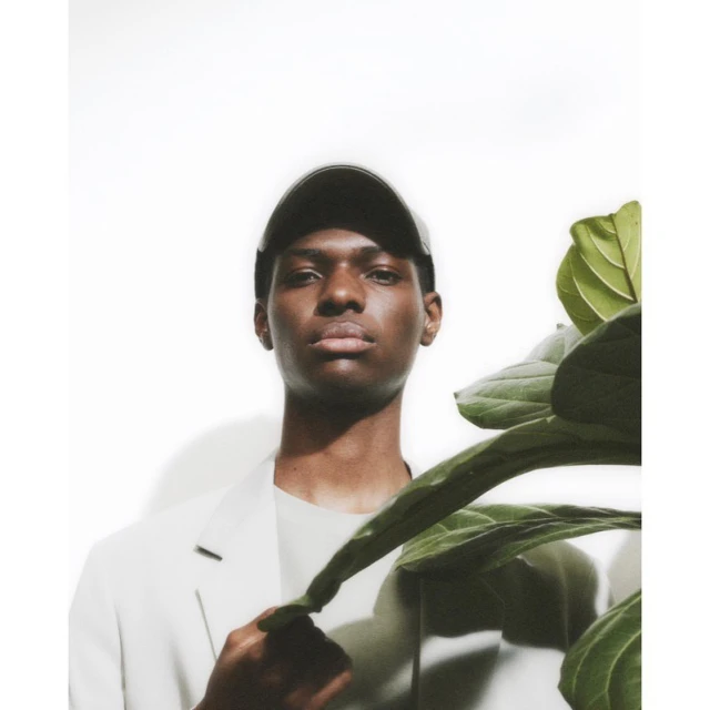 Portrait of a Black man wearing a baseball cap and blazer standing next to a fiddle-leaf fig plant in a brightly lit photo studio.