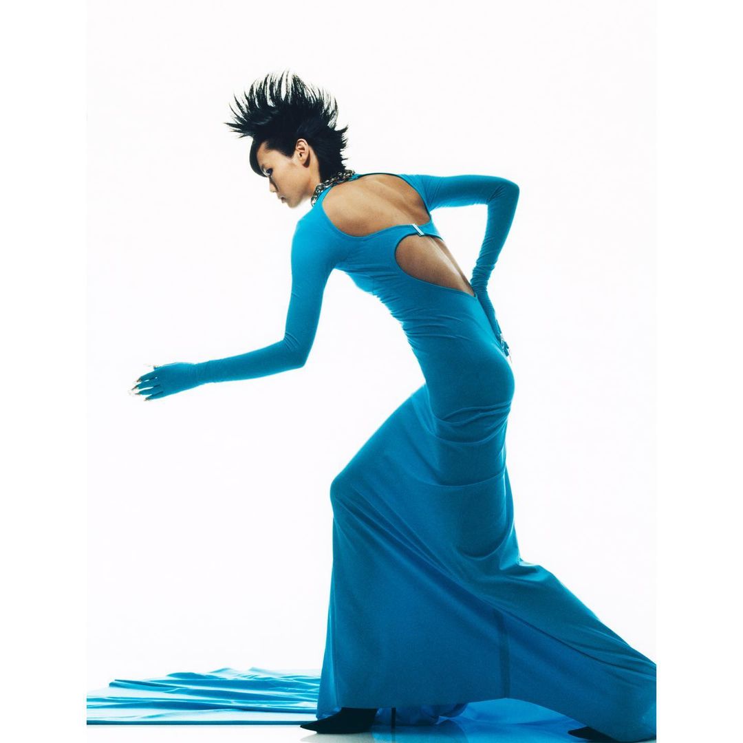 A woman in a long turquoise gown poses with outstretched hand against a bright white background in a fashion forward studio shot.