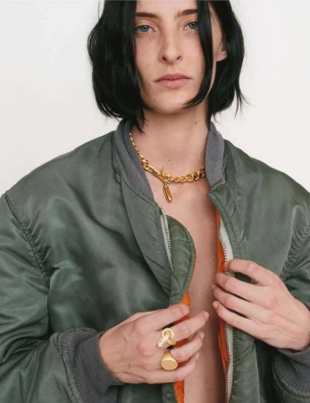 Model in olive green bomber jacket wearing gold chain necklace and statement rings against a white backdrop