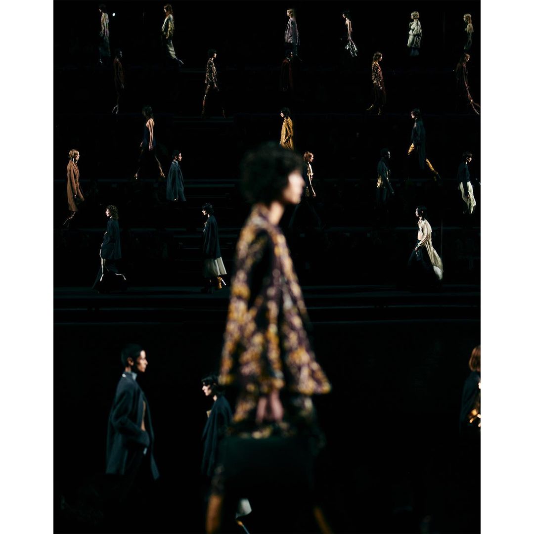 Runway show features several models walking in dark clothing during a fashion show, shallow depth of field blurs the foreground figure.