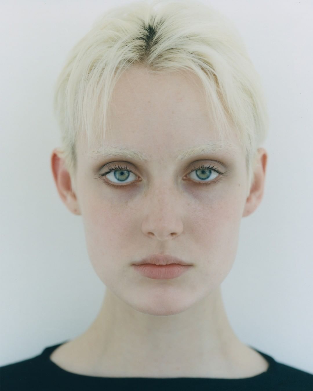 Close-up portrait of a fair-skinned woman with short blonde hair and light blue eyes, wearing a dark-colored top against a plain white background.