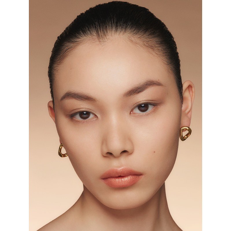Close-up portrait of a beautiful Asian woman with slicked-back hair, natural makeup, and gold earrings in front of a beige background.