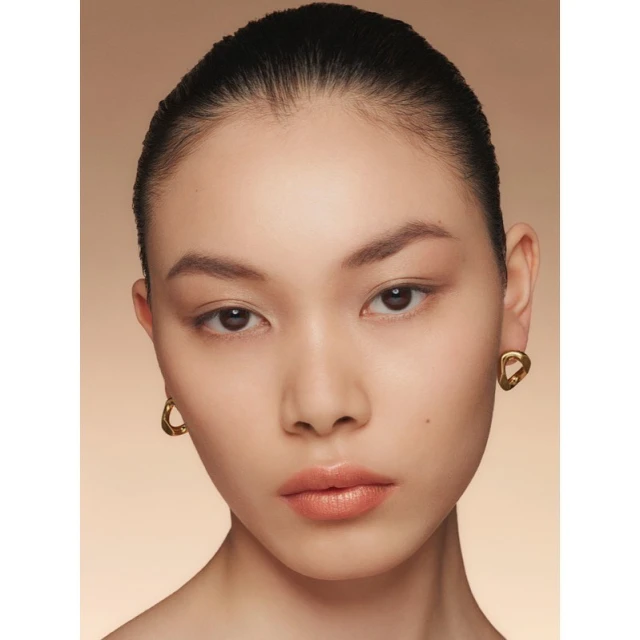 Close-up portrait of a beautiful Asian woman with slicked-back hair, natural makeup, and gold earrings in front of a beige background.