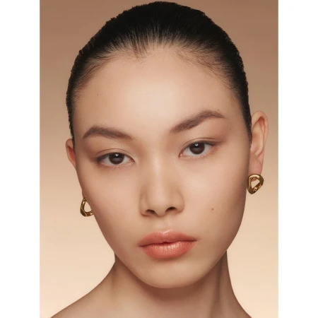 Close-up portrait of a beautiful Asian woman with slicked-back hair, natural makeup, and gold earrings in front of a beige background.