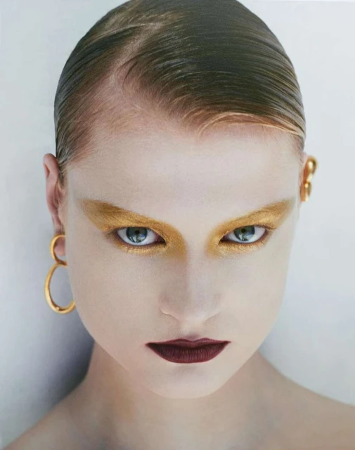 Close-up portrait of a woman with slicked-back hair, dramatic gold eyeshadow, dark lipstick, and gold earrings against a neutral backdrop.