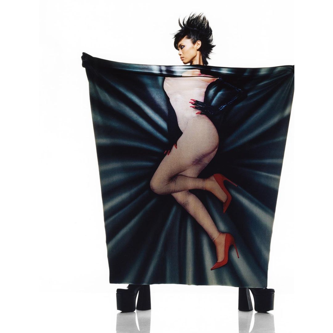 Artistic shot of a slender woman posing inside a draped print of herself, wearing red heels, against a bright white backdrop.