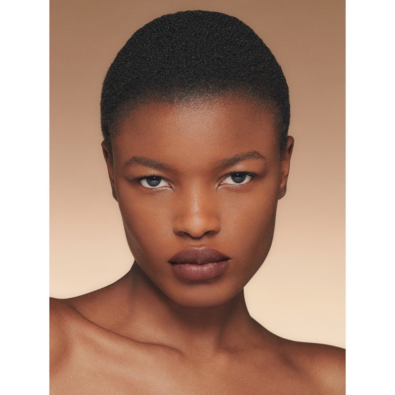 Close-up portrait of a Black woman with a bare shoulder against a warm, light-brown background, showcasing beauty and natural skin.