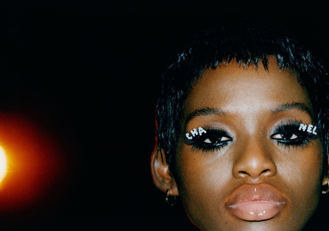 Close-up of a woman with decorative eyelashes spelling Chanel; black background and warm light source on the left side of the frame.