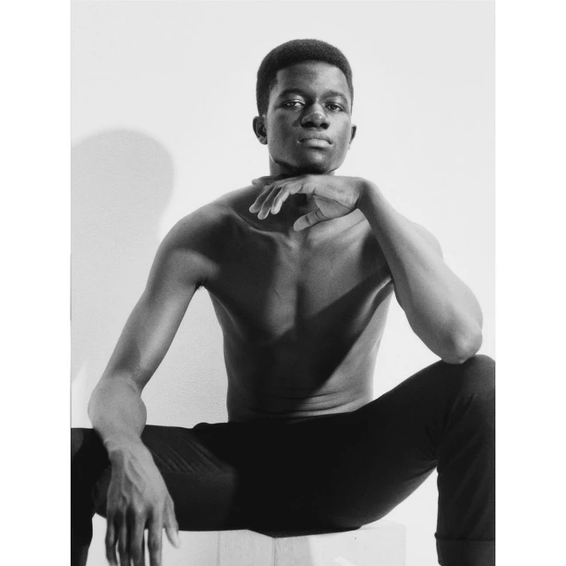 Black and white portrait of a shirtless man with dark hair and pants sitting on a white block in a photo studio with neutral background.