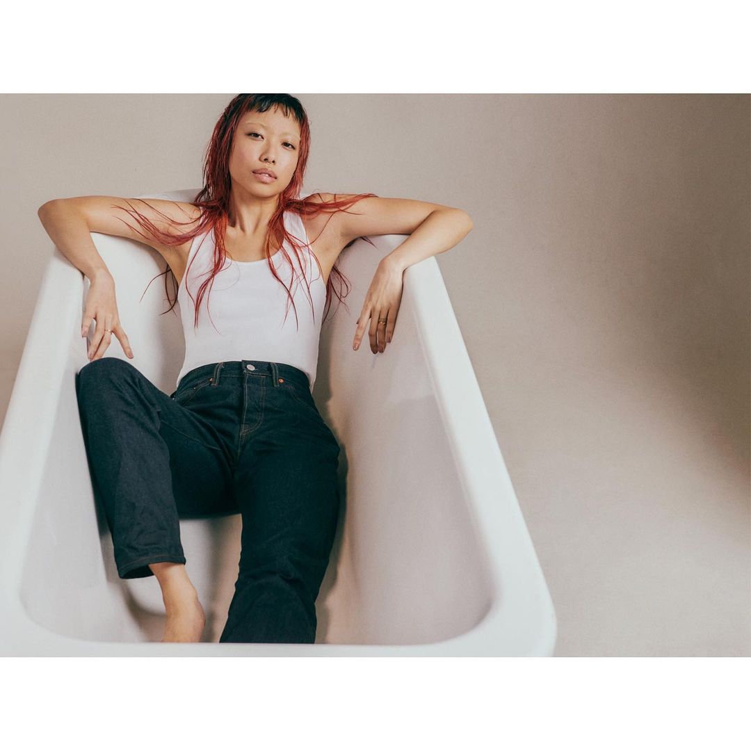 An adult woman with reddish hair poses in a white bathtub wearing a white tank top and dark jeans against a neutral background.