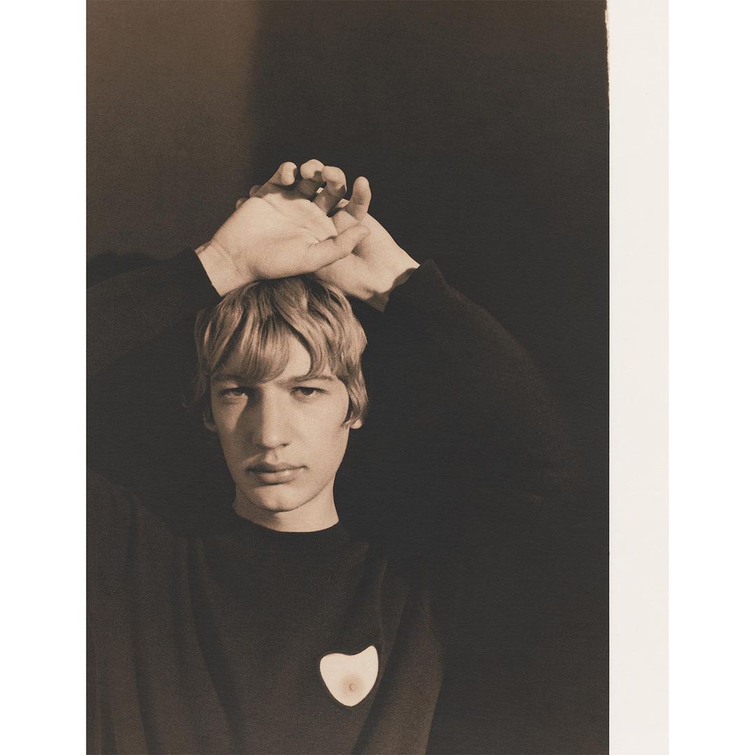 Portrait of a young adult with hands above their head in a dark sweater against a shadowed black backdrop in a sepia-toned photograph.