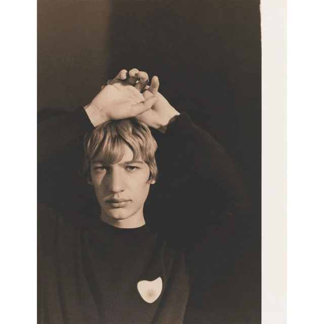 Portrait of a young adult with hands above their head in a dark sweater against a shadowed black backdrop in a sepia-toned photograph.