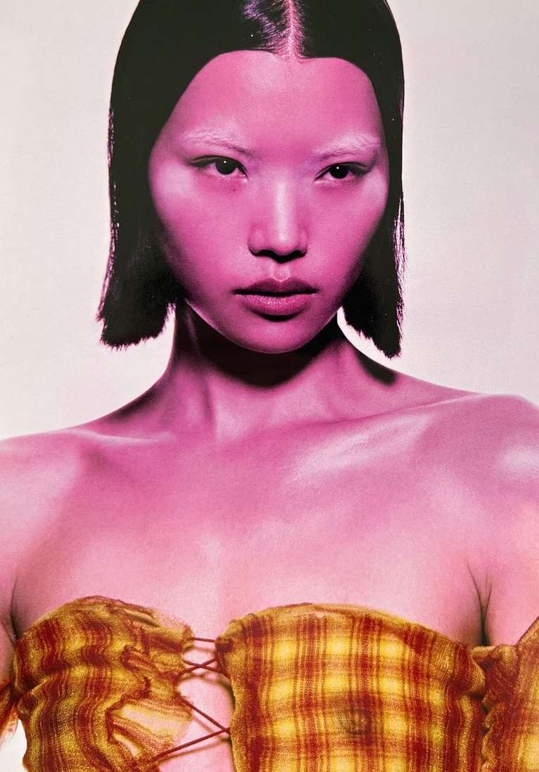 Eye-level portrait of an Asian female with short black hair, wearing a yellow plaid top, and pink skin in a light studio setting.