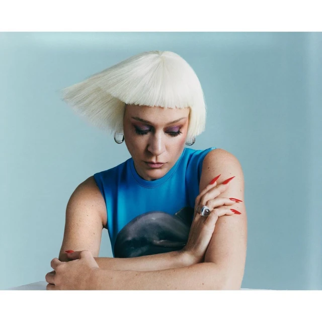 Portrait of a woman with a platinum bob haircut and bright makeup, wearing a blue top with a dolphin graphic, and long red nails.