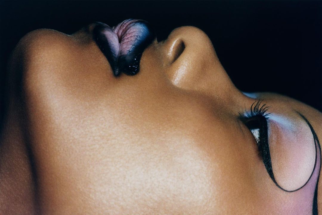 Close-up studio shot of a light-skinned adult female with dramatic black and blue makeup on her lips and eye in a dark studio setting.