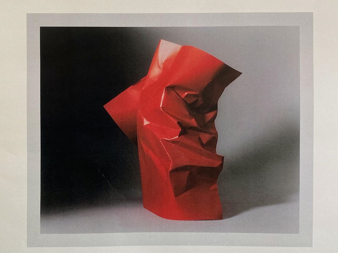 Abstract red paper sculpture with geometric folds, set against a gradient background, emphasizing texture and form in a studio setting.
