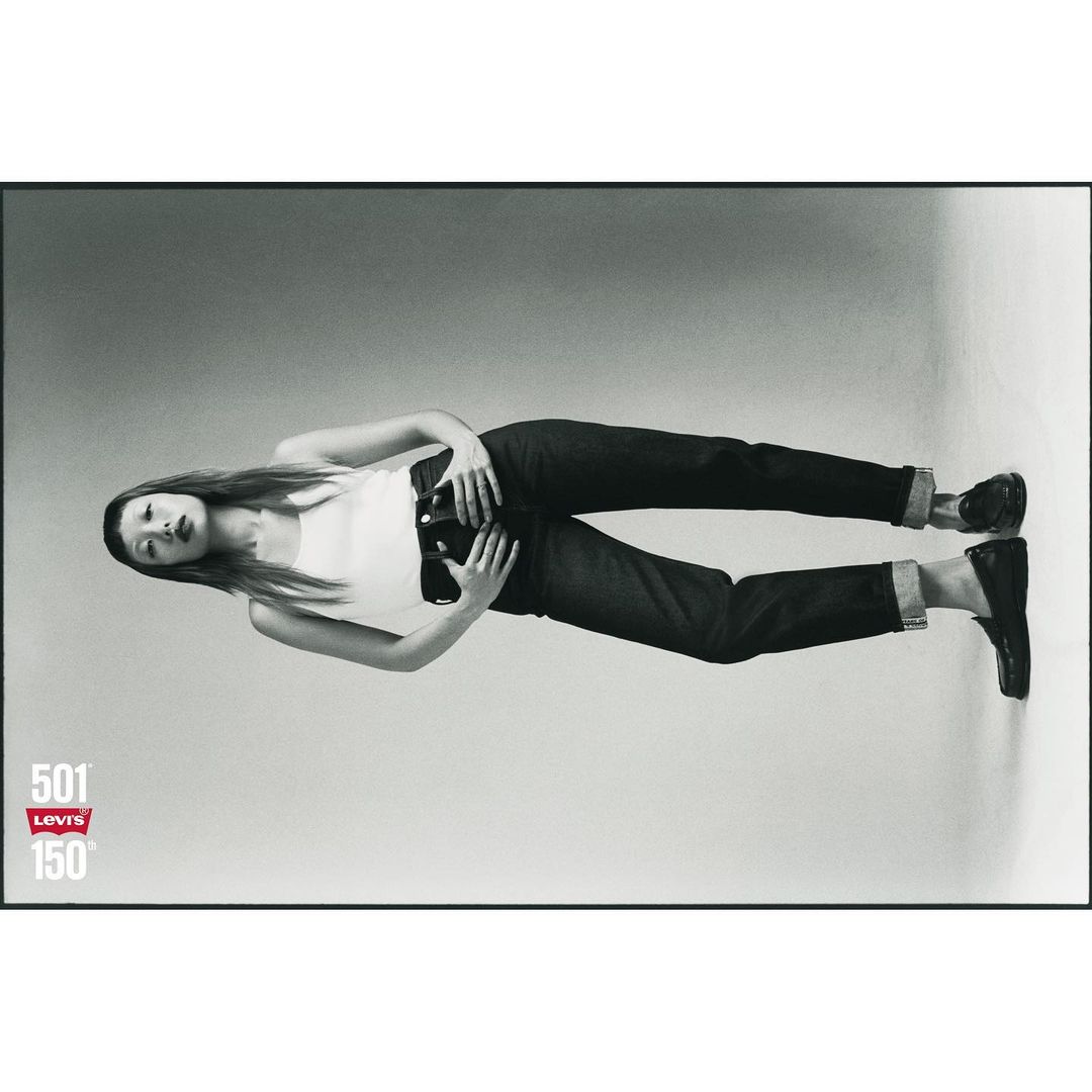 A woman lies on her side wearing blue jeans and a white tank top in a Levi's brand ad celebrating 150 years, studio shot on grey background.