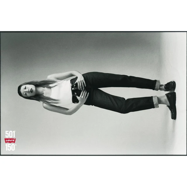 A woman lies on her side wearing blue jeans and a white tank top in a Levi's brand ad celebrating 150 years, studio shot on grey background.