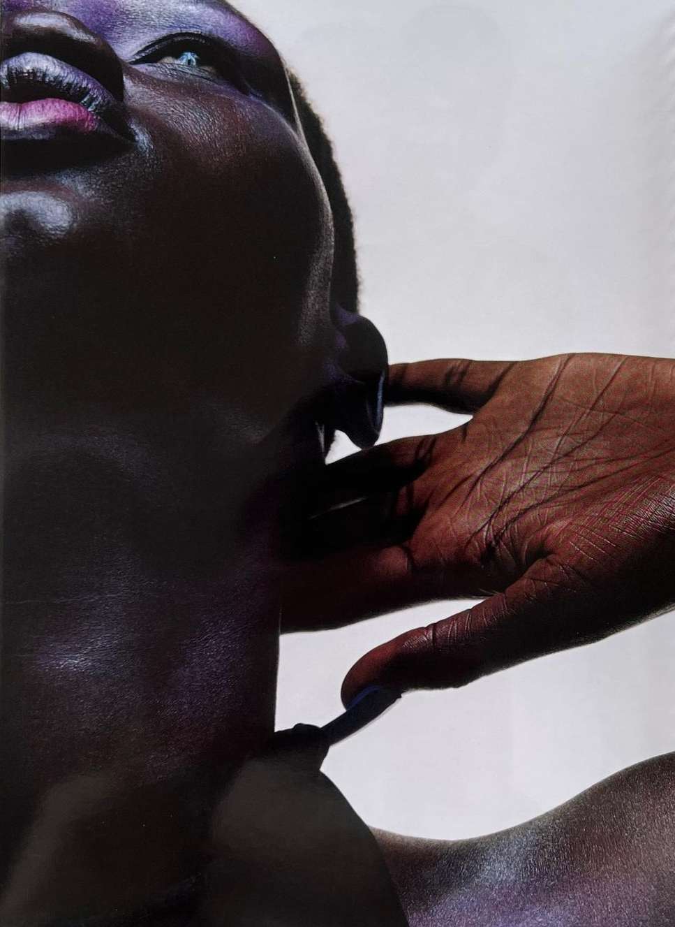 Close-up shot of a dark-skinned adult woman's face with vibrant purple eye makeup and glossy lips, her hand near her face.