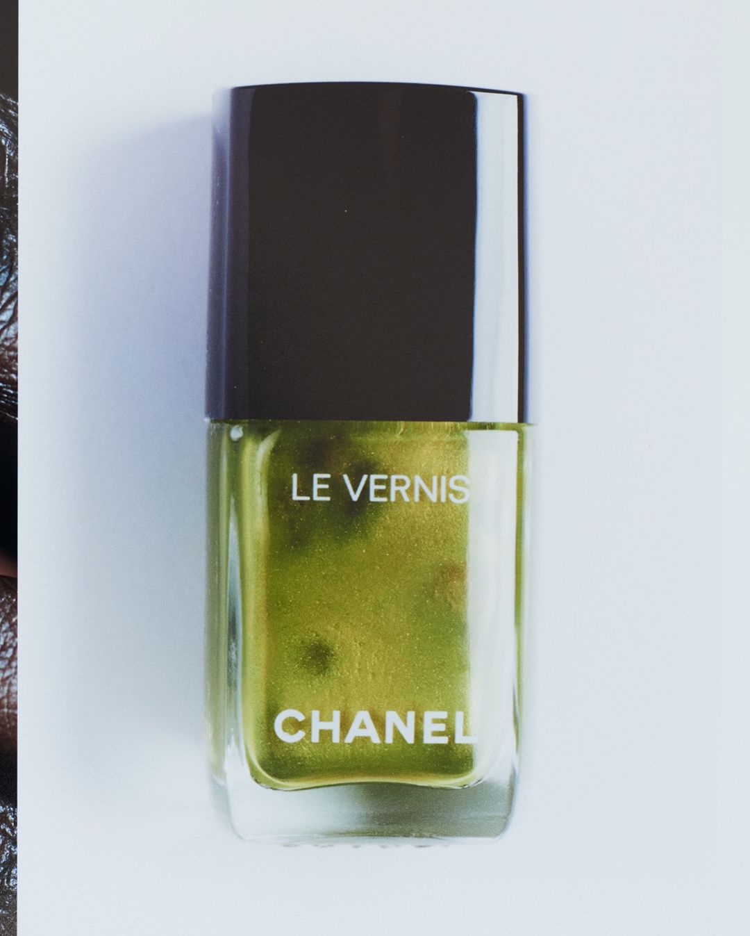 Close-up studio shot of a Chanel Le Vernis nail polish bottle with gold-green liquid, against a white background. Beauty product.