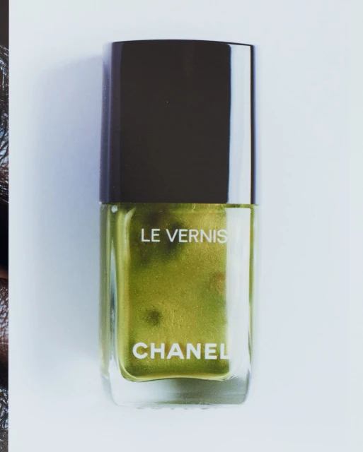 Close-up studio shot of a Chanel Le Vernis nail polish bottle with gold-green liquid, against a white background. Beauty product.