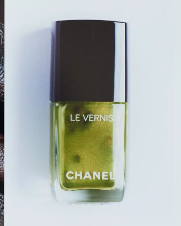 Close-up studio shot of a Chanel Le Vernis nail polish bottle with gold-green liquid, against a white background. Beauty product.