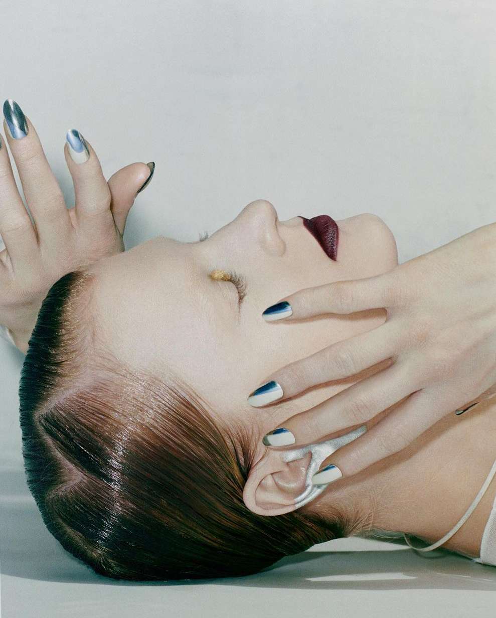 Close-up of a woman's face with metallic nail polish and dark lipstick in a beauty studio setting on a white background.