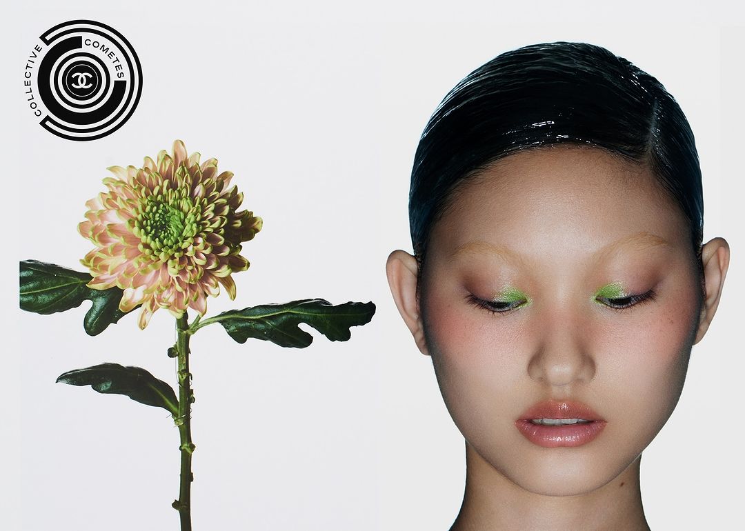 Close-up of a woman with green eyeshadow and slicked-back hair next to a light pink and green flower against a white background.