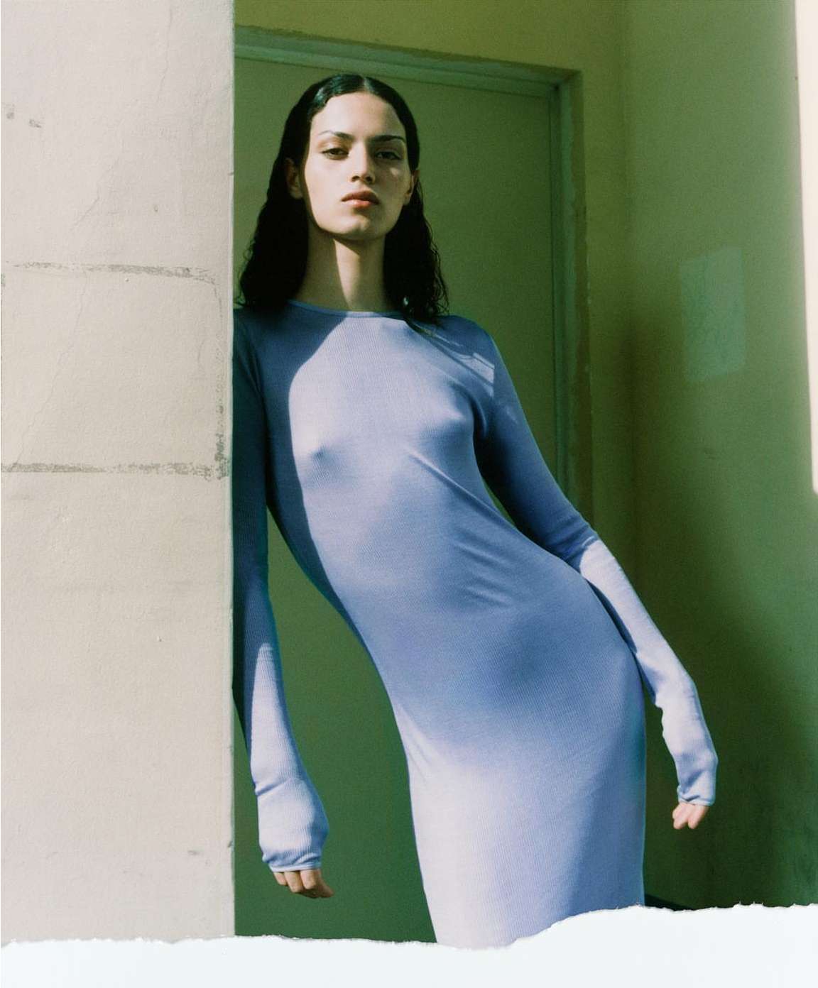 An adult female poses in a light blue dress against a textured wall with a green door, exhibiting fashion and editorial style.