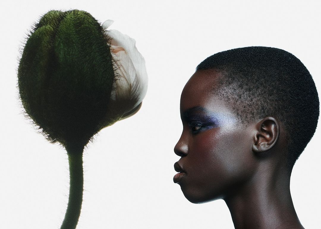 Profile portrait of a short-haired dark-skinned adult woman with iridescent blue makeup next to a large flower bud against a white background.