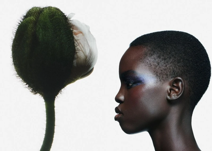 Profile portrait of a short-haired dark-skinned adult woman with iridescent blue makeup next to a large flower bud against a white background.