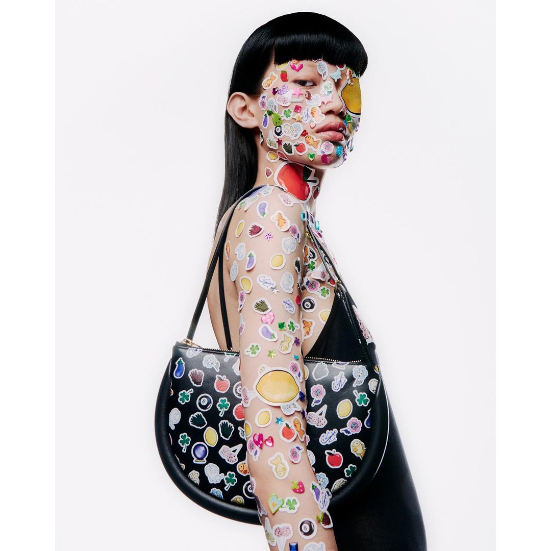 Woman in black top covered with fruit and emoji stickers, carrying black leather purse in a studio shot on a white backdrop.