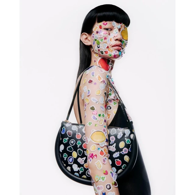 Woman in black top covered with fruit and emoji stickers, carrying black leather purse in a studio shot on a white backdrop.