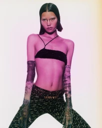 A female model wearing a black crop top, patterned trousers with a gold Versace belt, and sheer gloves against a gradient background.