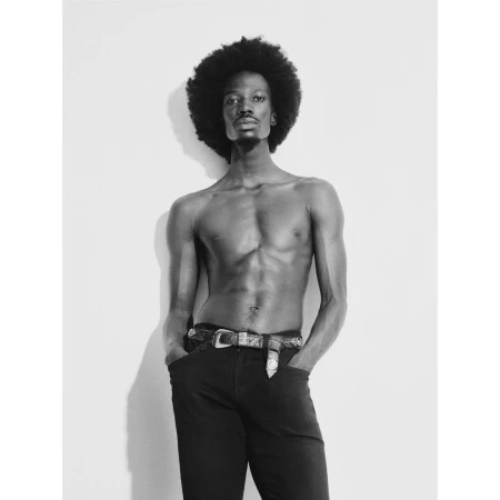 Black and white studio portrait of a shirtless man with an afro hairstyle wearing dark trousers and a decorated belt.