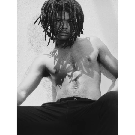 Monochrome portrait of a shirtless man with dreadlocks seated against a plain textured wall in New York.