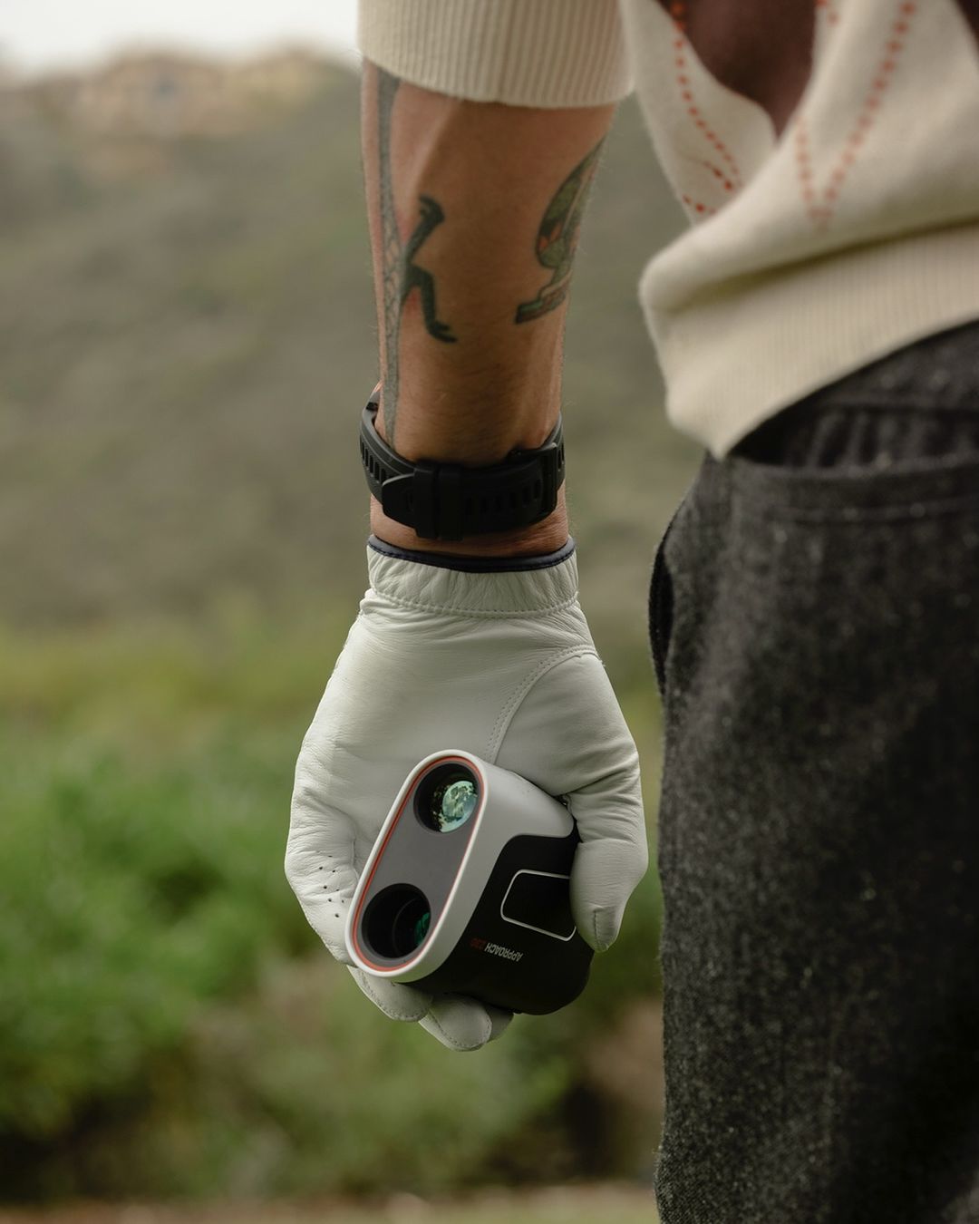 A gloved hand holds a golf rangefinder in a close up shot on a golf course with green grass and trees in the background.