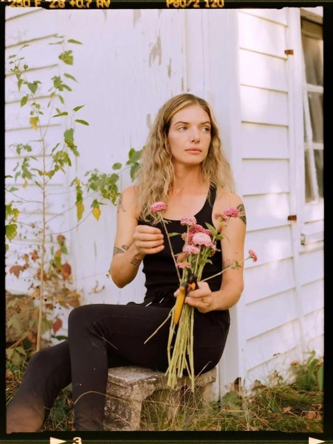 Woman in black tank top sits outdoors arranging pink flowers, with a white rustic building and greenery in the background, natural light.