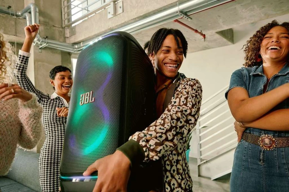A group of smiling young adults celebrate while a man holds a lit-up JBL PartyBox speaker indoors, creating a lively party atmosphere.