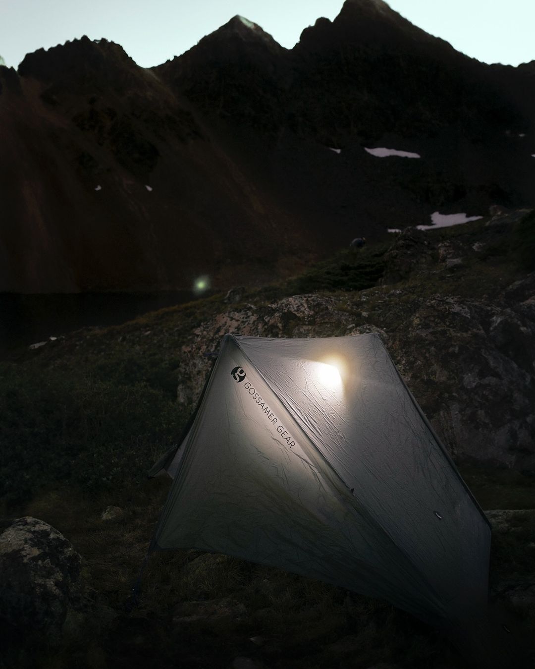 Illuminated tent at dusk in a mountainous landscape with a lake, camping at twilight under a dark sky with visible mountain peaks.