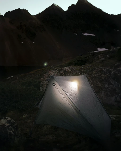Illuminated tent at dusk in a mountainous landscape with a lake, camping at twilight under a dark sky with visible mountain peaks.