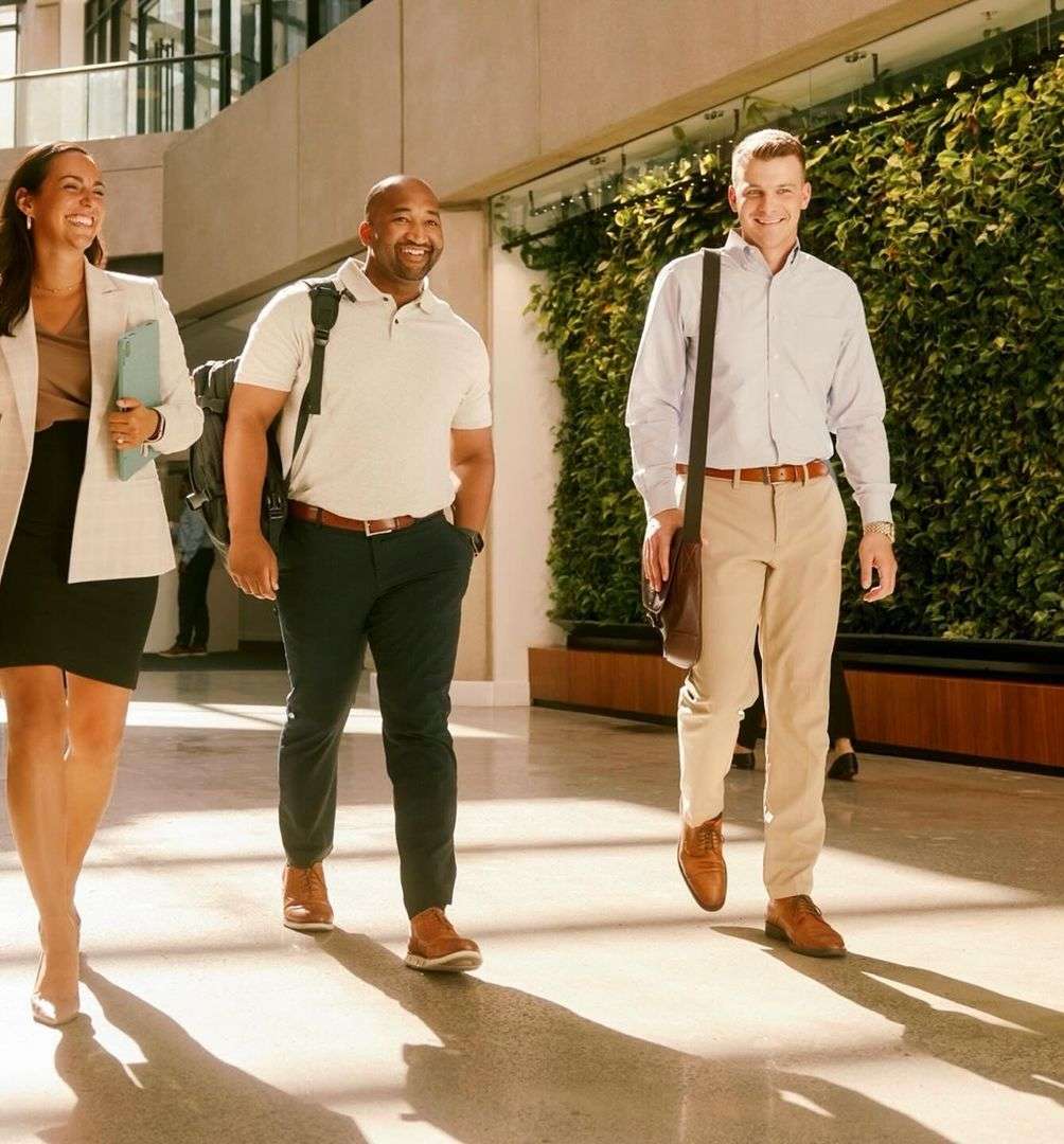 Three smiling professionals—a woman and two men—walk through a bright, modern office lobby with a living wall, bathed in warm light.