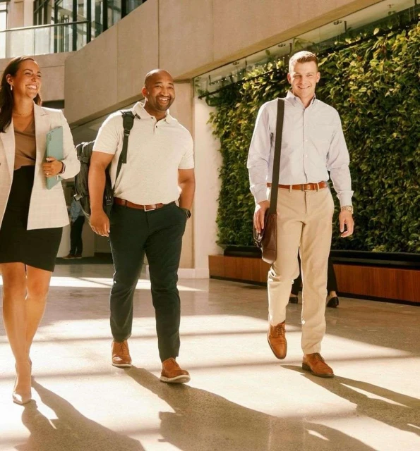 Three smiling professionals—a woman and two men—walk through a bright, modern office lobby with a living wall, bathed in warm light.