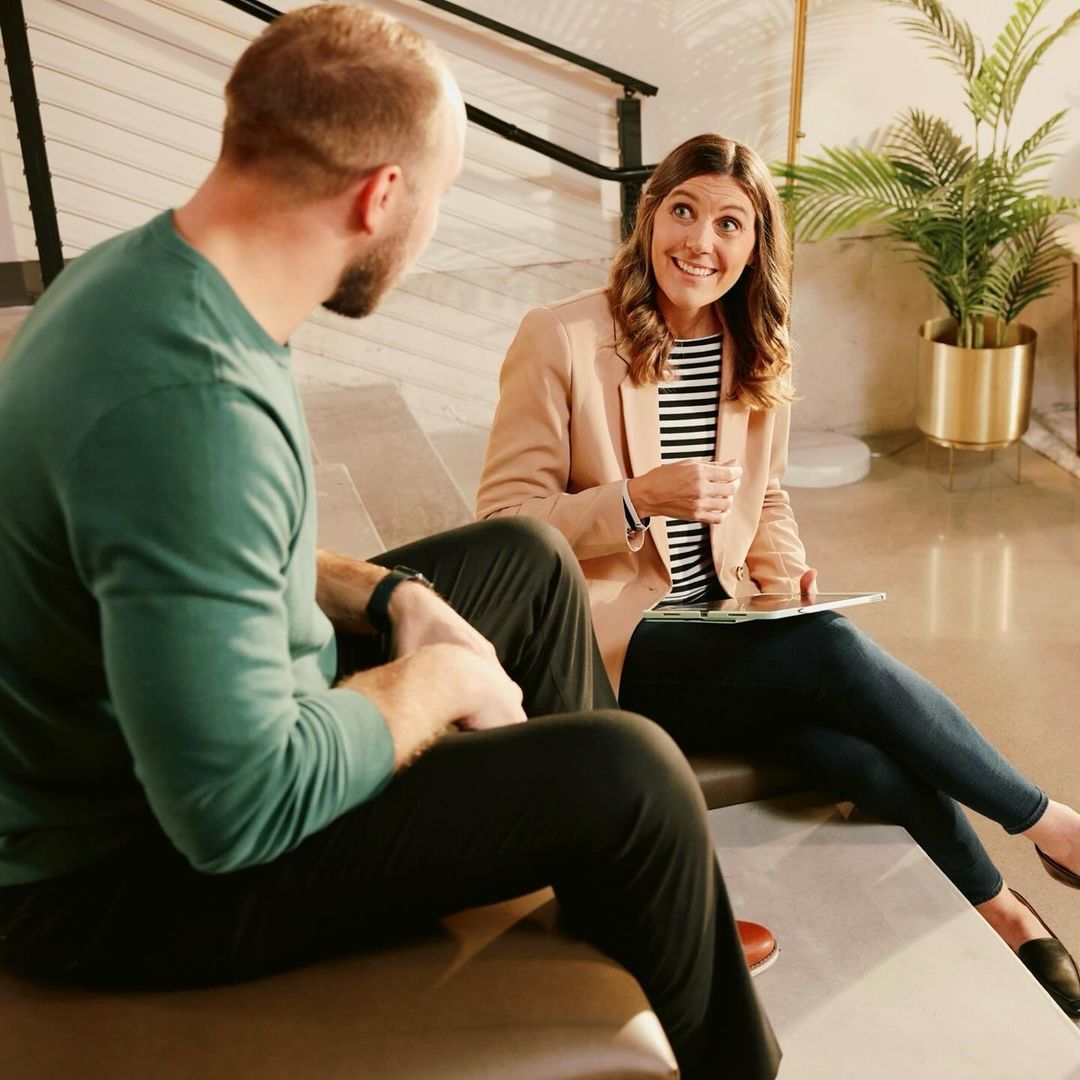 Two people sitting indoors: an adult man in green and an adult woman in a blazer, smiling and holding a tablet, indoor setting.