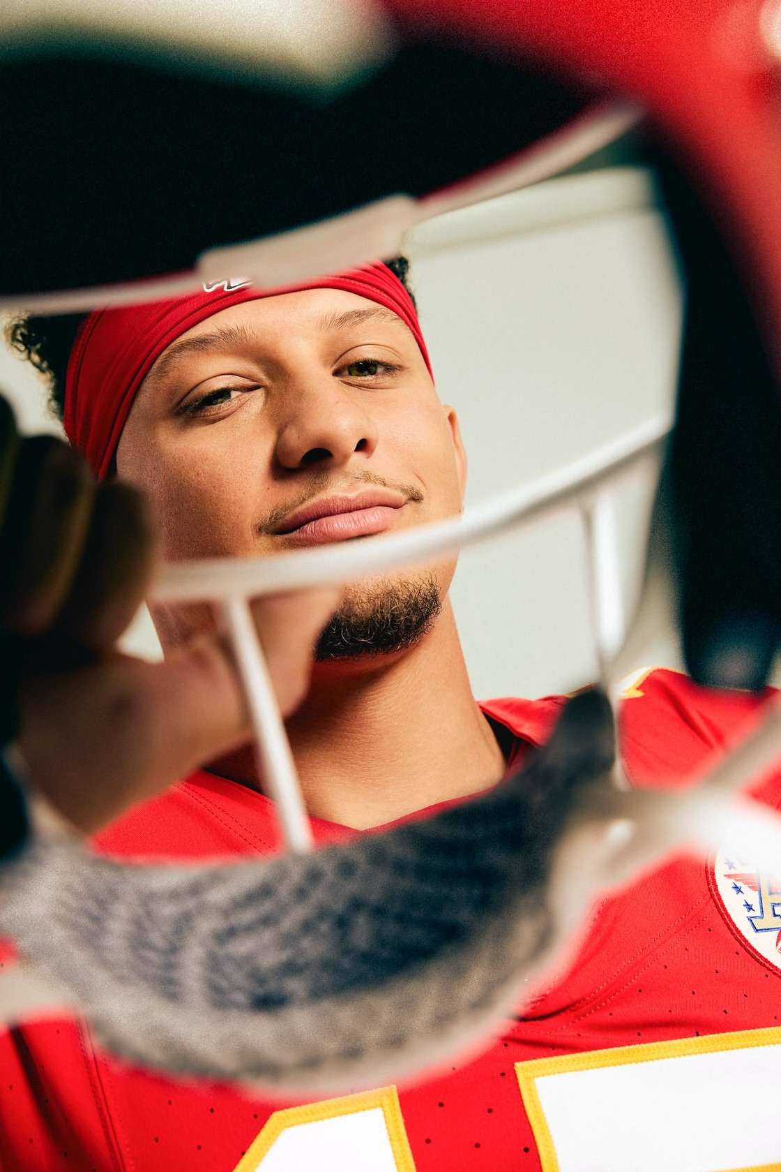 Close up of a male American football player with a red headband and jersey, looking through the bars of a white helmet.