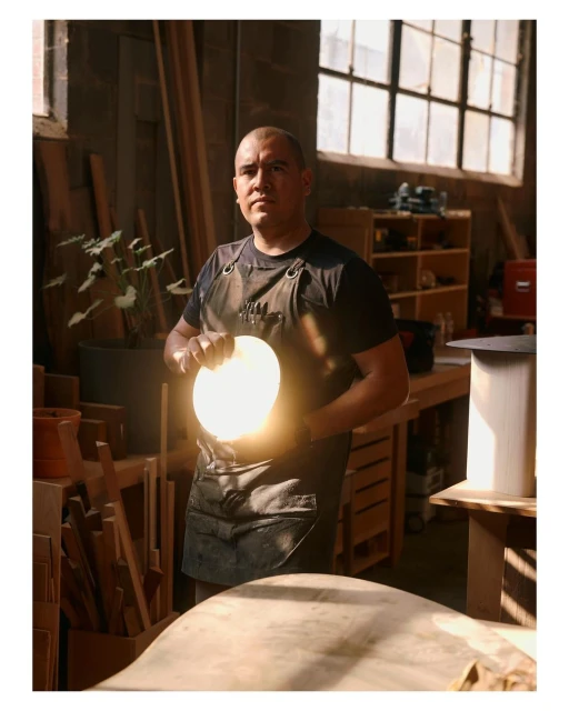 Adult male artisan holds a glowing circular object in his workshop with wooden materials, tools, and natural light from a window.