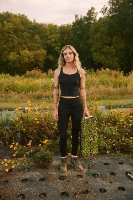 Woman in black tank top stands among yellow flowers in a garden holding plants on a farm with rows of greenery and trees.