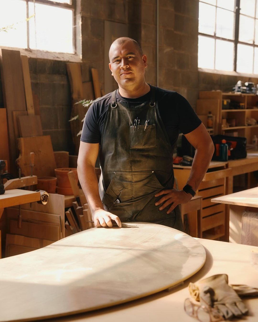 Adult male woodworker in a workshop setting, leaning on a large, light-colored wooden board in a close-up, medium shot.